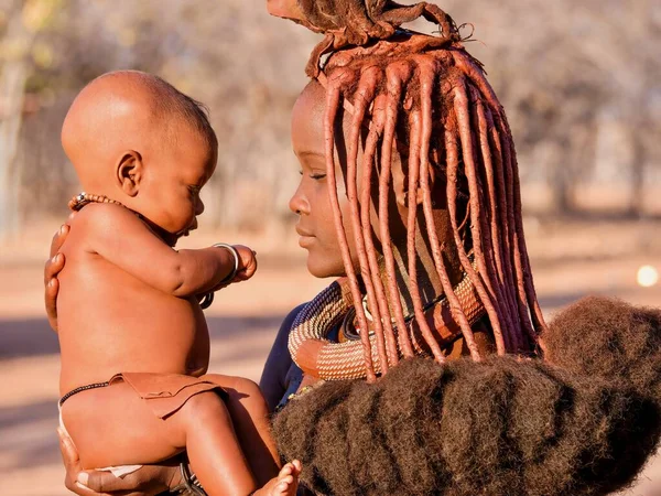 Palmwag, Namibya - 21 Ağustos 2016. Genç bir Afrikalı kadın, Himba kabilesinin geleneksel saç stili ve krem rengi deri macununu sürerken bebeğini tutuyor..