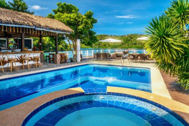 Filipinler 'in Puerto Galerisi' nde, güneşli bir günde, canlı mavi suyu olan güzel tropik bir tatil köyü yüzme havuzu ve suyun kenarında sazdan bir çatısı olan açık hava barı..