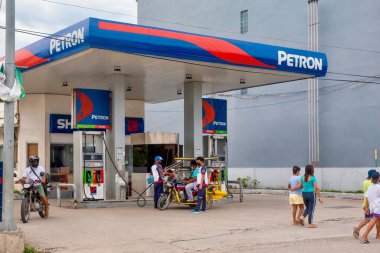 Puerto Galera, Philippines - May 21, 2021. Street view of a Petron service station, where a tricycle is being filled with gasoline by an attendant wearing a Covid mask. clipart