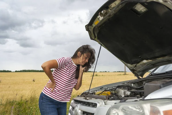 A broken car. A young woman stands on the road by a broken car against the background of a yellow field. The hood is open. I need help. Helplessly looks under the hood. A phone call to help with a broken car.