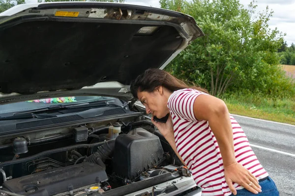 A broken car. A young woman stands on the road by a broken car against the background of a yellow field. The hood is open. I need help. Helplessly looks under the hood. A phone call to help with a broken car.