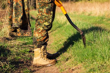 Kamuflajlı bir turistin bacakları istihkamcı küreğiyle doğa üzerindeki parlak yeşil çimen yürüyüş konseptinin etrafındaki patikada duruyor.