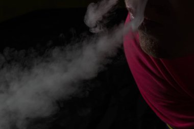 Sigara ve uyuşturucu bağımlılığı gibi karanlık bir arka plan kavramına karşı ağzındaki gri buharı çıkaran bir insanın yüzünün bir parçası.