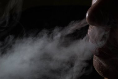 Çerçevenin kenarında sigara içen bir insanın yüzü ağızdan ve burundan kötü alışkanlık ve elektronik sigara konseptinden yoğun gri buhar salıyor.