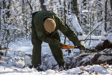 Askeri üniformalı bir adam istihkamcı küreğiyle çukur kazıyor. Kış ormanında tek başına her tarafı beyaz karla kaplamış.