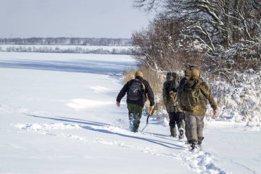 Kamuflajlı gezginler parlak bir kış gününde karla kaplı bir arazide yürüyorlar.
