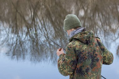 Balıkçı elinde bir kaşık tutuyor, nehrin bulanık arka planında balık tutmadan önce yemi kontrol ediyor.