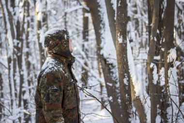 Askeri üniformalı bir adam kış ormanlarında beyaz karlarla kaplı bir şekilde profilde görünür.