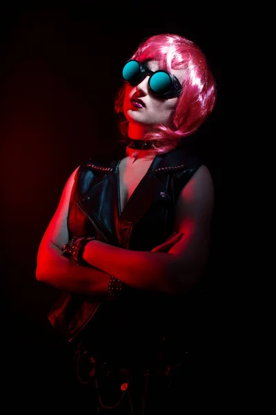 Pembe saçlı, dudak delen, deri yelek giyen, yakalı ve kolları çapraz gözlüklü havalı genç bir rockçı kadın..
