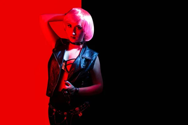 Young beautiful rocker girl with pink hair and lip piercing wearing leather vest and spiked collar on red and black background.