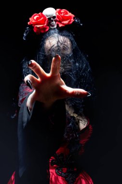Kızıl gotik elbiseli cadı ya da büyücü kadın, siyah peçeli, kafatası ve güllerle kaplı taç karanlıktan elini uzatıyor. Cadılar Bayramı konsepti.