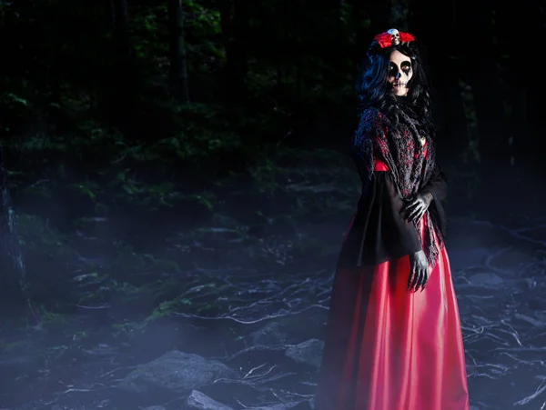 夜霧の森の中に立って頭蓋骨やバラと赤いゴシックドレス ショールや王冠でサンタ ムエルテの化粧を持つ神秘的な女性 ハロウィンのコンセプト — ストック写真