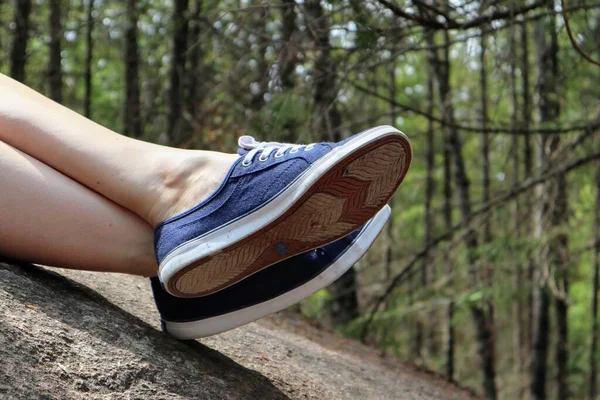 Close-up of a woman\'s crossed legs in blue sneakers lie on a stone. Forest in the background