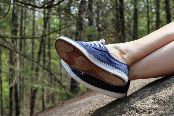 Close-up of a woman\'s crossed legs in blue sneakers lie on a stone. Forest in the background