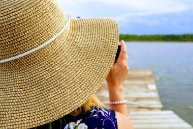 Bir kadın şapkası ve bir kadının omzunun kenarına yakın çekim. Bayan fotoğraf çekiyor. Arka planda göl, orman ve ahşap iskele var..