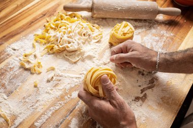 Erkek eller ahşap masada makarna için hamuru hazırlıyor, etrafında merdane, un ve tagliatelle var.