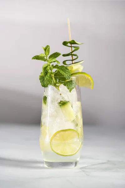 A tall long drink glass with cucumber, mint, ice cubes and lime on a white background