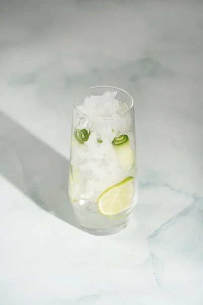 A tall long drink glass with cucumber, ice cubes and lime on a white background