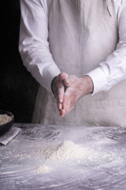 Beyaz gömlekli bir erkek aşçı ve gri bir pron siyah bir mutfak masasında un ile el çırpıyor.