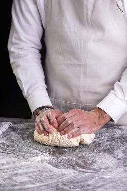 Beyaz gömlekli, gri önlüklü, siyah mutfak masasında hamur yoğuran bir aşçı.