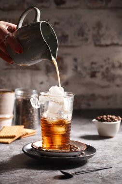 Kadın eli, buzlu ve kahveli bir bardağa süt döküyor.