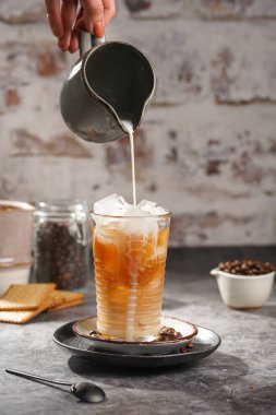 Gri bir kavanozdan, içinde buz küpleri ve kahve olan bir bardağa süt dökmek.