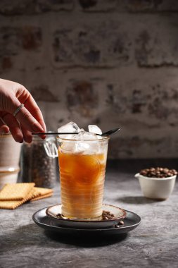 Kadın eli, buzlu ve kahveli bir bardağa siyah çay kaşığı koyuyor.