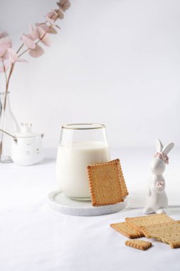 Mermer bir tabakta bir bardak süt beyaz bir masa örtüsü, taze pişmiş kurabiyeler, ve beyaz paskalya tavşanları, heykelcikler.