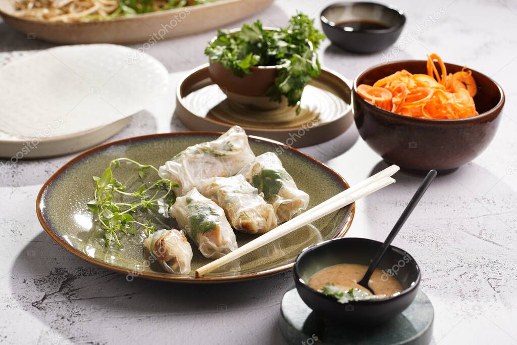 Several spring rolls with chicken, carrot and bamboo sprouts in rice paper on green plate, sesame sauce and coriander on concrete surface