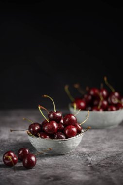 Tatlı ve ekşi kırmızı kirazların üzerine siyah arka planda iki gri skandal kasesi serpiştiriyor.