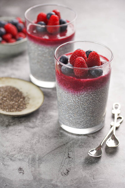 Two glasses with chia seeds pudding, raspberry jam and fresh raspberries and blueberries on a grey surface