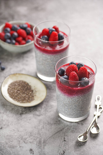 Two glasses with chia seeds pudding, raspberry jam and fresh raspberries and blueberries on a grey surface