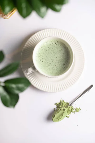 A white porcelain cup with japanese matcha tea drink on a white saucer plate on a white surface, a tea spoon with matcha powder, bamboos whisker and a green plant zamioculcas , top view