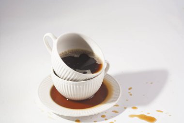 Beyaz bir yüzey üzerinde beyaz bir tabak üzerinde başka bir kupaya istiflenmiş kapuçino dolu beyaz bir porselen bardak.