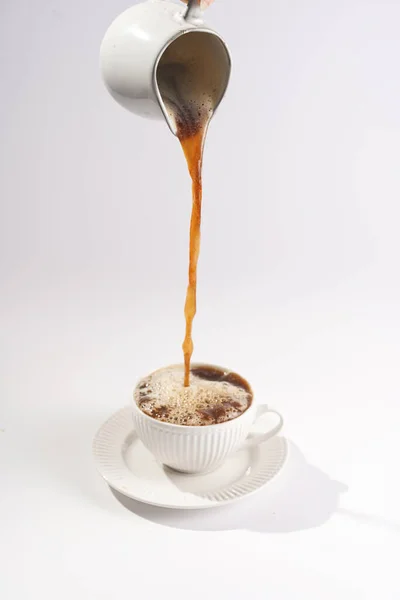 Pouring coffee into a white porcelain cup with coffee drink cappuccino on a white saucer plate on a white surface