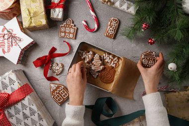 Noel süsü ve hediyelerle çevrili gri bir yüzeyde ev şeklinde beyaz şekerli zencefilli kurabiyeler ve kurabiyeli bir kutu kurabiye tutan kadın elleri.