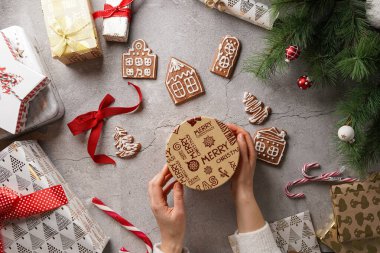 Beyaz şekerli zencefilli kurabiyeler. Gri bir yüzeyde ev şeklinde. Noel süslemeleri ve hediye tutan kadın elleri.