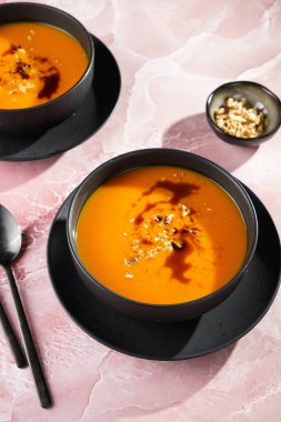 İki koyu siyah kase, turuncu kabak kremalı çorba, kabak tohumu yağı, iki siyah kaşık pembe mermer yüzey