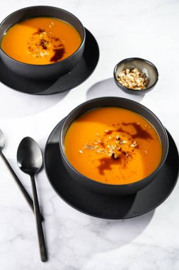 İki koyu siyah kase, turuncu kabak kremalı çorba, kabak tohumu yağı, iki siyah kaşık pembe mermer yüzey