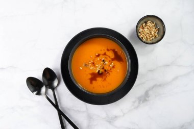 Kabak çekirdeği yağlı turuncu kabak kremalı bir kase, mermer bir yüzeyde iki siyah kaşık.