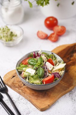 Küçük, beyaz bir salata kasesi içinde mikroyeşiller, kiraz domatesleri ve tahta bir tahtada taze fesleğen yaprakları gri beton bir yüzey üzerinde.