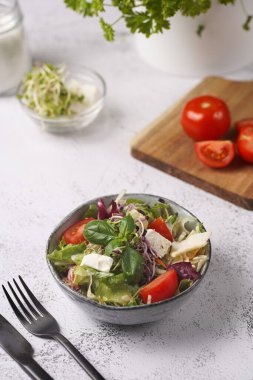 Küçük, beyaz bir salata kasesi içinde mikroyeşiller, kiraz domatesleri ve tahta bir tahtada taze fesleğen yaprakları gri beton bir yüzey üzerinde.