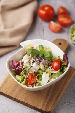 Küçük, beyaz bir salata kasesi içinde mikroyeşiller, kiraz domatesleri ve tahta bir tahtada taze fesleğen yaprakları gri beton bir yüzey üzerinde.