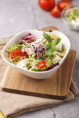 Küçük, beyaz bir salata kasesi içinde mikroyeşiller, kiraz domatesleri ve tahta bir tahtada taze fesleğen yaprakları gri beton bir yüzey üzerinde.