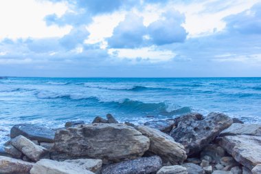 Alacakaranlıkta sakin Kıbrıs plajı, ön planda kayalar, sakin deniz ve huzurlu bir sahil havası yaratan serin mavi akşam tonları.