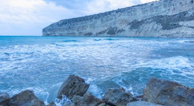 Alacakaranlıkta sakin Kıbrıs plajı, ön planda kayalar, sakin deniz ve huzurlu bir sahil havası yaratan serin mavi akşam tonları.