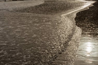 Akşam vakti Kıbrıs 'ta kumlu bir plajın yüzeyinde. Suyun üzerinde yumuşak altın yansımalı sakin sörf, huzurlu Akdeniz kıyısı atmosferi