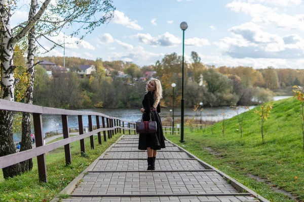 Eine schöne Frau steht und lehnt am Geländer, ein Erwachsener mit charismatischer Erscheinung in schwarzer Kleidung, im Herbst vor dem Hintergrund von Bäumen, blauer Himmel. — Stockfoto