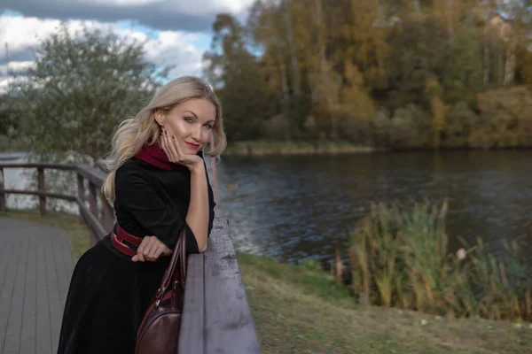 En vacker dam står och lutar sig på räcket, med vackra ögon i svarta kläder, på hösten mot en bakgrund av blå moln. — Stockfoto