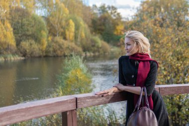 Bir bayan ayakta duruyor ve korkuluklara yaslanıyor, siyah elbiseli narin dudakları olan bir yetişkin, sonbaharda ağaçların arka planında mavi bulutlara yaslanıyor..